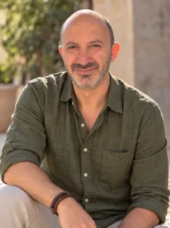 Raul Ortegon-Bolio, sonriendo, con camisa verde olivo de mangas arremangadas, sentado al aire libre bajo el sol con plantas y un muro de piedra detrás.